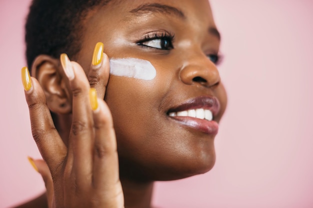 Close-upportret van het gezicht van een Afrikaanse vrouw die het aanbrengen van creme op het gezicht toepast en dagelijkse routine-isolatie doet...