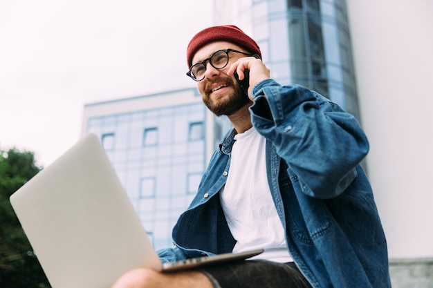 Foto close-upportret van moderne gebaarde mannelijke hipster in oogglazen die op telefoon spreken en tegelijkertijd laptop met behulp van.