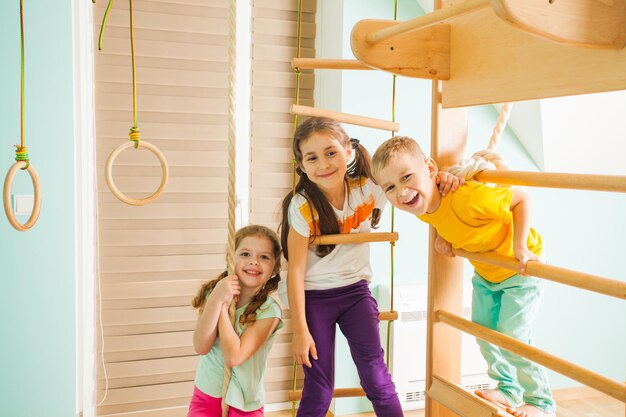 Close view portrait of two adorable girls and a cute boy, all smiling and hanging on a swedish wall and rope-ladder. Active kids spending their leisure time training, holding, looking at the camera