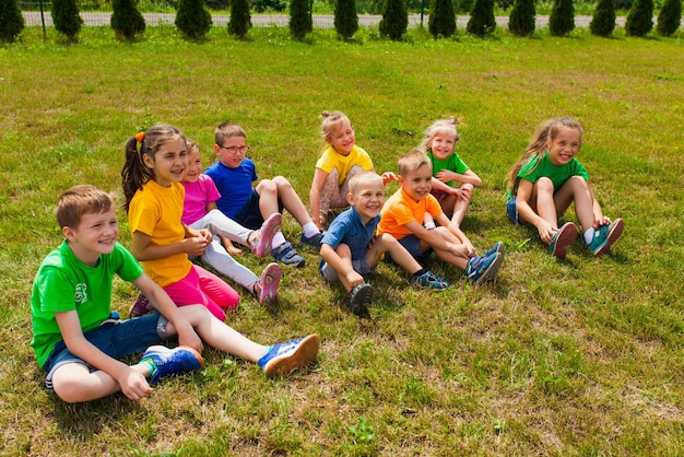 녹색 잔디에 앉아 화려한 티셔츠를 입은 다양한 연령대의 웃고 있는 아이들의 모습을 가까이서 볼 수 있습니다. 방과 후 여름 가족 캠프 참가자들은 풀밭에 앉아 웃고 웃고 있다