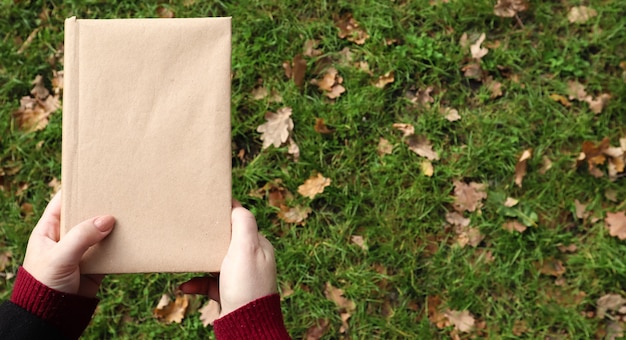 A closed book in a cover made of craft paper in female hands with green grass and fallen yellow leaves in the background. Flat lay, top view. Template, layout. Copy space.