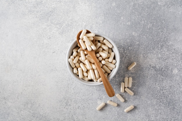 Closed collagen in capsules and a wooden spoon for diet food in a ceramic bowl on an old gray concrete table. Flat lay with copy space.