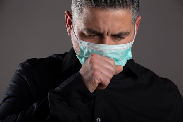 Closed portrait of man with surgical mask coughing in studio on gray background, healthcare concept.