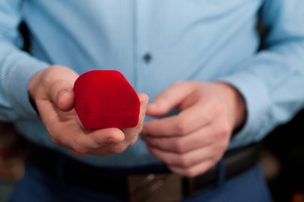 A closed red velvet ring box in the hands of a man in a blue shirt