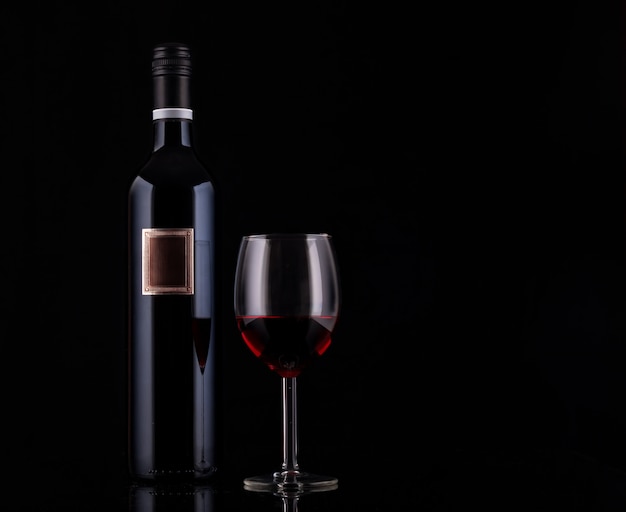 Closed red wine bottle with empty label and glass of wine on black background with reflections