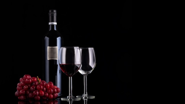 Closed red wine bottle with empty label, grape and two glasses on black background with reflections and copy space