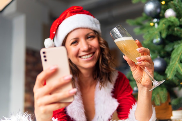 아름다운 여성 모자와 빨간 산타클로스가 행복하게 웃고 있고, 샴페인 잔, 컨셉의 휴일 축하, 크리스마스 트리 배경을 닫았습니다. 크리스마스 인사말 온라인. 새해 자가격리.