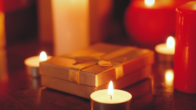 Closeup 4k footage of golden gift box and burning candles on wooden table at night. Perfect background or backdrop for Christmas or New Year