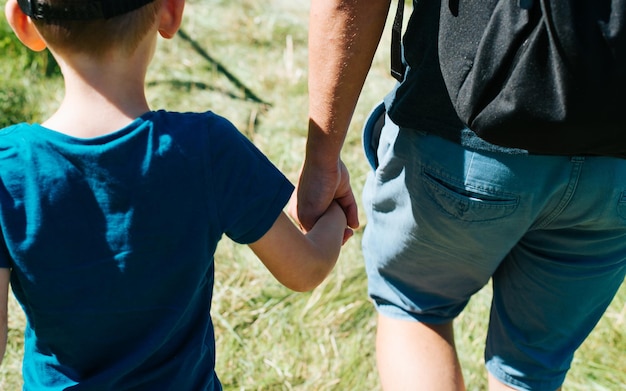 Closeup of an adult man and little boy holding hands together outdoors Rear view of dad and son walking in nature Selective focus on hands