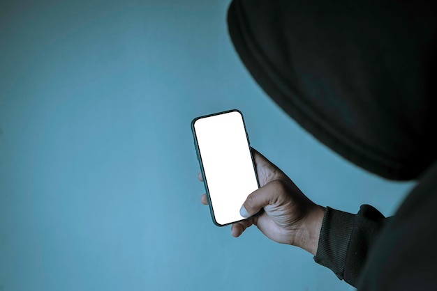 CloseUp An anonymous hacker wearing a mask and a black hoodie shows off a smartphone with a blank white screen Hacking and Malware Concepts