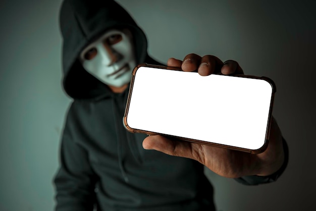 Closeup an anonymous hacker wearing a mask and a black hoodie\
shows off a smartphone with a blank white screen hacking and\
malware concepts