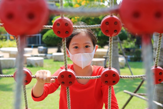 Bambina asiatica del primo piano che indossa una maschera protettiva mentre gioca sulla rete della corda di arrampicata all'aperto