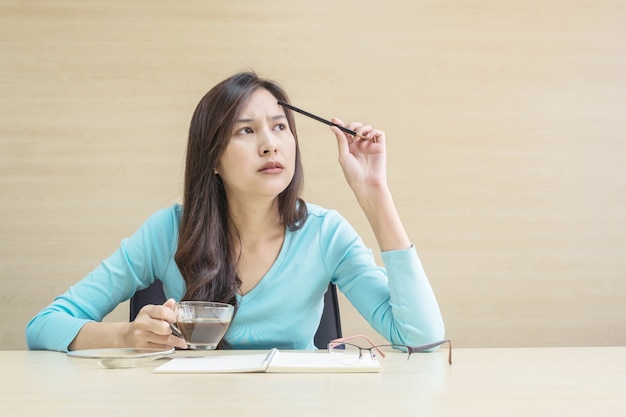 Женщина крупного плана азиатская работая с думая лицом и карандашем и чашкой кофе