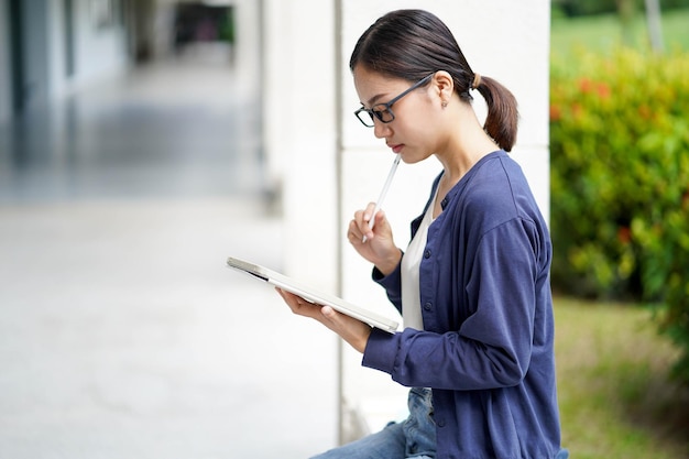 アジア系女子生徒がカジュアルな服を着て読書をし校舎の試験用のタブレットに短いメモを書き背景がぼやけています アジア系学校のコンセプト