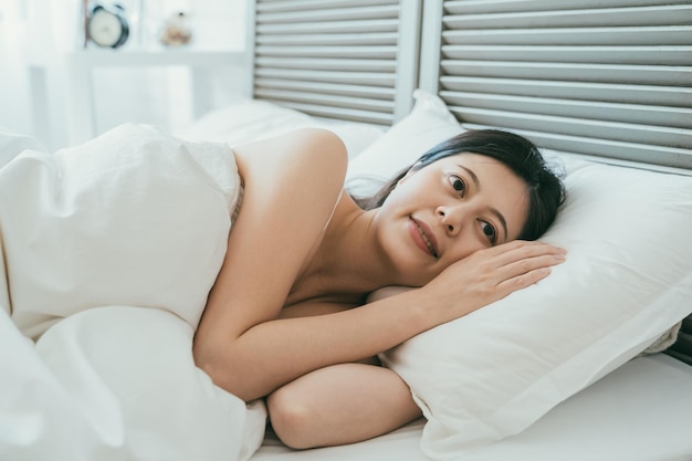 Closeup attractive asian woman lying awake on her side in bed\
is smiling feeling happy for getting a good sleep at a bright\
comfortable home bedroom interior.