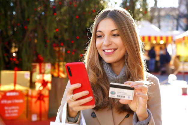 魅力的な若い女性のクローズ アップ彼女のプレゼントをスマート フォンでオンラインで支払い、クリスマス マーケットとクレジット カードと背景にボックスとツリー