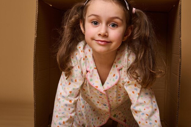 Closeup baby girl with two ponytails in pajamas peeking out from behind a cardboard box isolated over beige background with copy space Home housing insurance and real estate concept