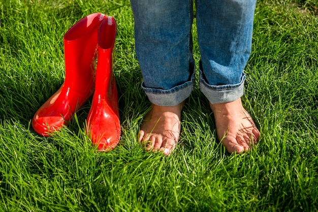 Closeup barefoot girl standing next red garden gumboots on grass