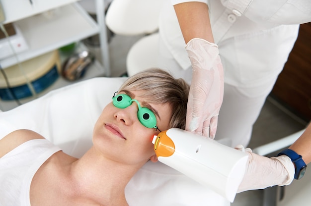 Closeup of beautician doing laser rejuvenation in a beauty salon. Elos epilation hair removal procedure on young woman face. Facial skin care. Hardware ipl cosmetology