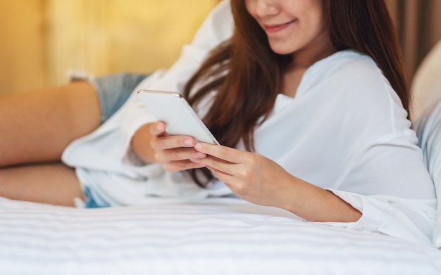 Closeup  of a beautiful asian woman using and looking at mobile phone while lying down on a white bed