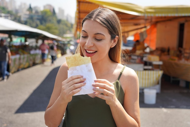 Photo closeup of beautiful girl eating brazilian traditional food pastel de feira in market fair of sao paulo brazil