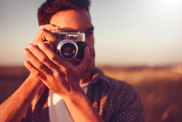 Foto primo piano bellissimo ritratto di giovane uomo bello con fotocamera vintage scattare una foto sullo sfondo del campo e sul tramonto umore estivo autunnale fotografia