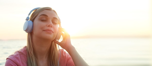 Closeup beautiful woman in the sunset sunlight listening to music in blue headphones with her eyes c