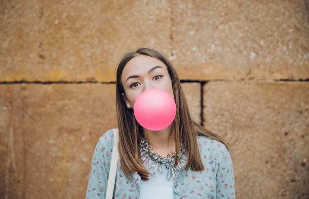 Foto primo piano di bella giovane ragazza bruna che soffia gomma da masticare rosa su un muro di pietra stone