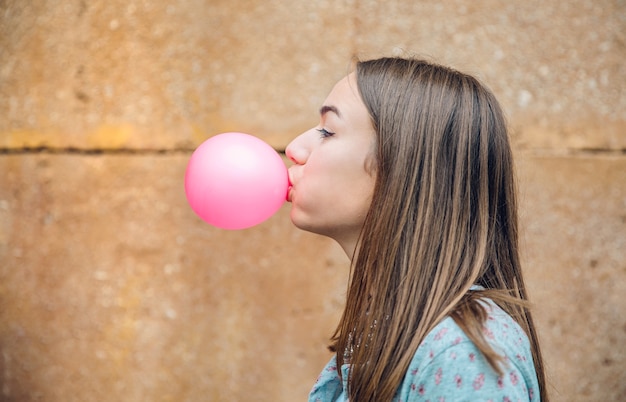 Closeup of beautiful young brunette teenage girl blowing pink bubble gum over a stone wall