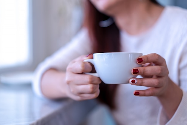 Closeup beauty portrait model hands with red fashion nails painting in warm sweater holding a white cup of coffee