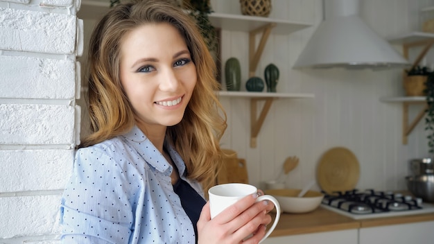 Closeup blond girl with loose flowing hair drinks tea smiles leaning on wall against shelves with kitchen utensils