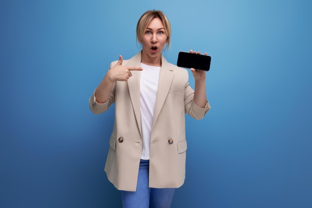 Closeup of blonde millennial woman in jacket showing smartphone with mockup on studio background