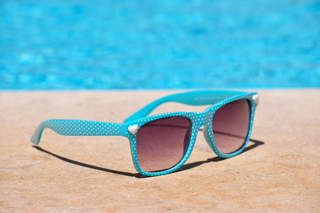 Closeup of blue sunglasses on swimming pool side at tropical resort on warm sunny day. Summer vacation concept.