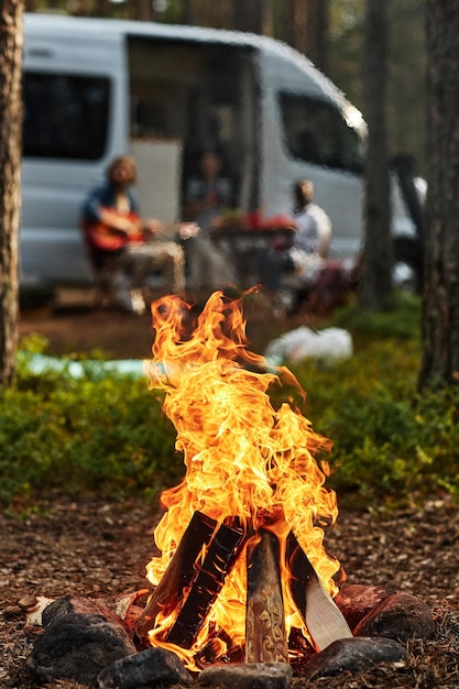 Closeup of bonfire during camp in the forest with young people playing guitar in background