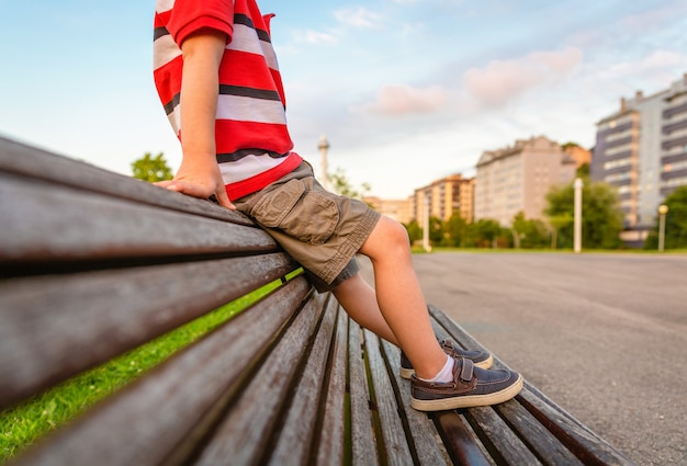 Closeup of boy legs with short pants sitting on the top of wooden bench park relaxing in a bored summer day