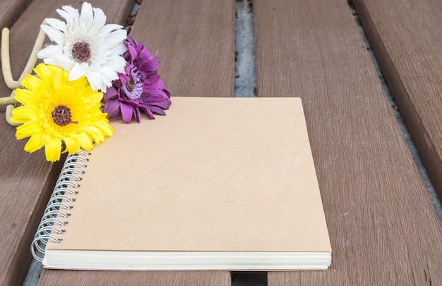 Closeup brown note book with colorful faked flower on blurred old wood table textured background