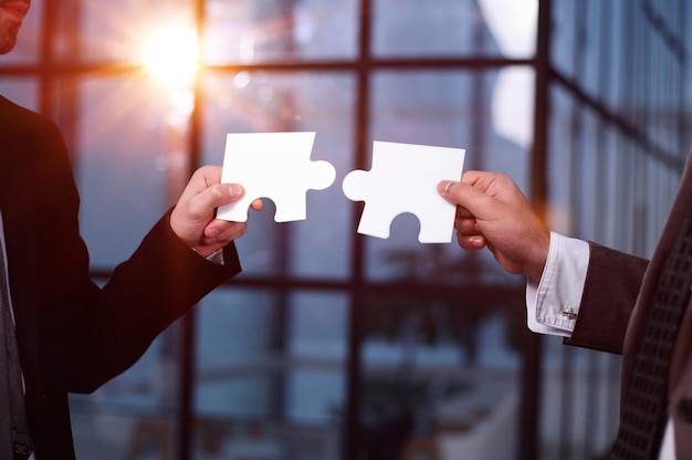 Closeup of business partners putting together two white puzzle pieces against the backdrop of an office building