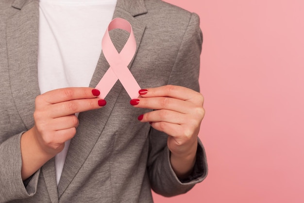 乳がんのリスクと社会意識の女性の健康保険医療保険屋内スタジオのピンクのリボンの警告を保持しているエレガントなスーツのジャケットのビジネスパーソンのクローズアップはピンクの背景を撮影しました