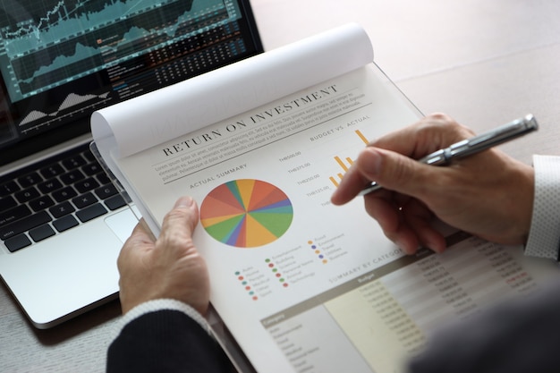 Closeup on businessman or analyst hands holding financial statements reviewing on return on investment, ROI, investment risk analysis in front of laptop computer with graphs and related information