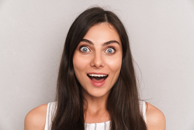 Foto primo piano di una donna caucasica eccitata con lunghi capelli scuri che sorride ed esprime sorpresa isolata su un muro grigio