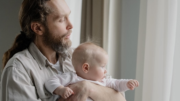 Primo piano padre caucasico barbuto papà che tiene bambino figlia figlio neonato neonato guardando fuori dalla finestra