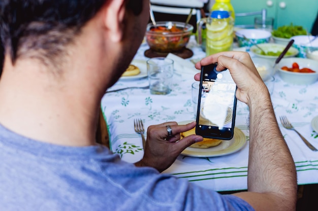 テーブルで提供される食べ物の写真を撮る彼の携帯電話で座っている白人男性のクローズアップ