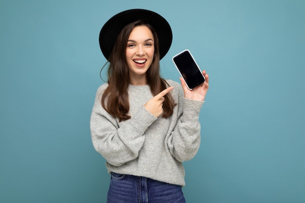 Closeup of charming young happy woman wearing black hat and grey sweater holding phone looking at