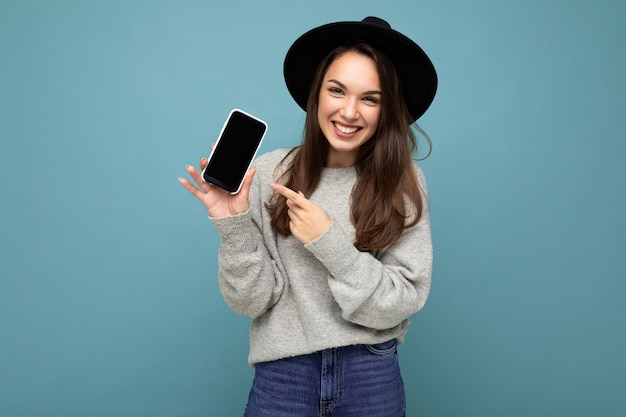 Closeup of charming young happy woman wearing black hat and grey sweater holding phone looking at