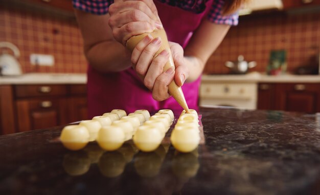 Foto primo piano dello chef in grembiule rosa spremitura di cioccolato bianco fuso con liquido cremoso dolce dal sacchetto di pasticceria in stampi per caramelle raccolte per preparare tartufi di cioccolato fatti a mano