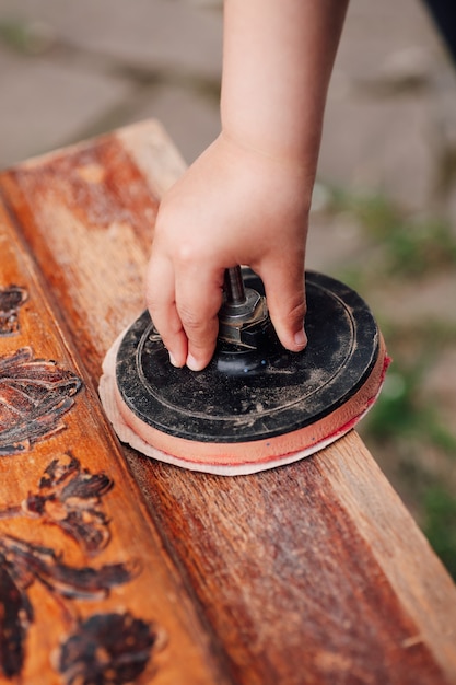 Closeup a childs hand holds a roll with sandpaper and scours an old wooden table with a floral woode...