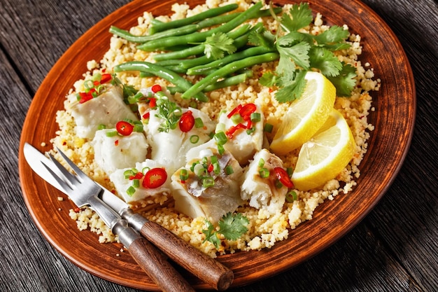 Closeup of cod with bulgur steamed green beans sprinkled with hot chili peppers and spring onions on a rustic plate on a dark wooden table balkan cuisine