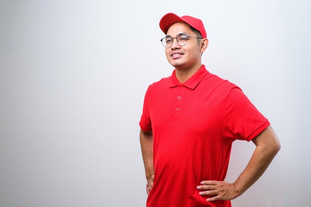 Closeup of confident smiling asian male courier in red uniform and cap smiling determined hand on hip