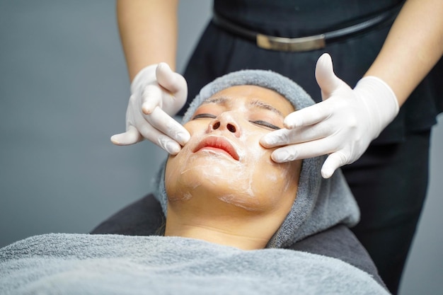 Closeup cosmetologist applying smooth facial white mask cream and massage on beauty woman's face