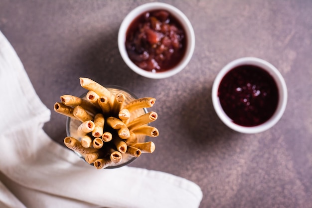 Closeup of crispy sweet straws with berry filling and two bowls of jam in a container Top view